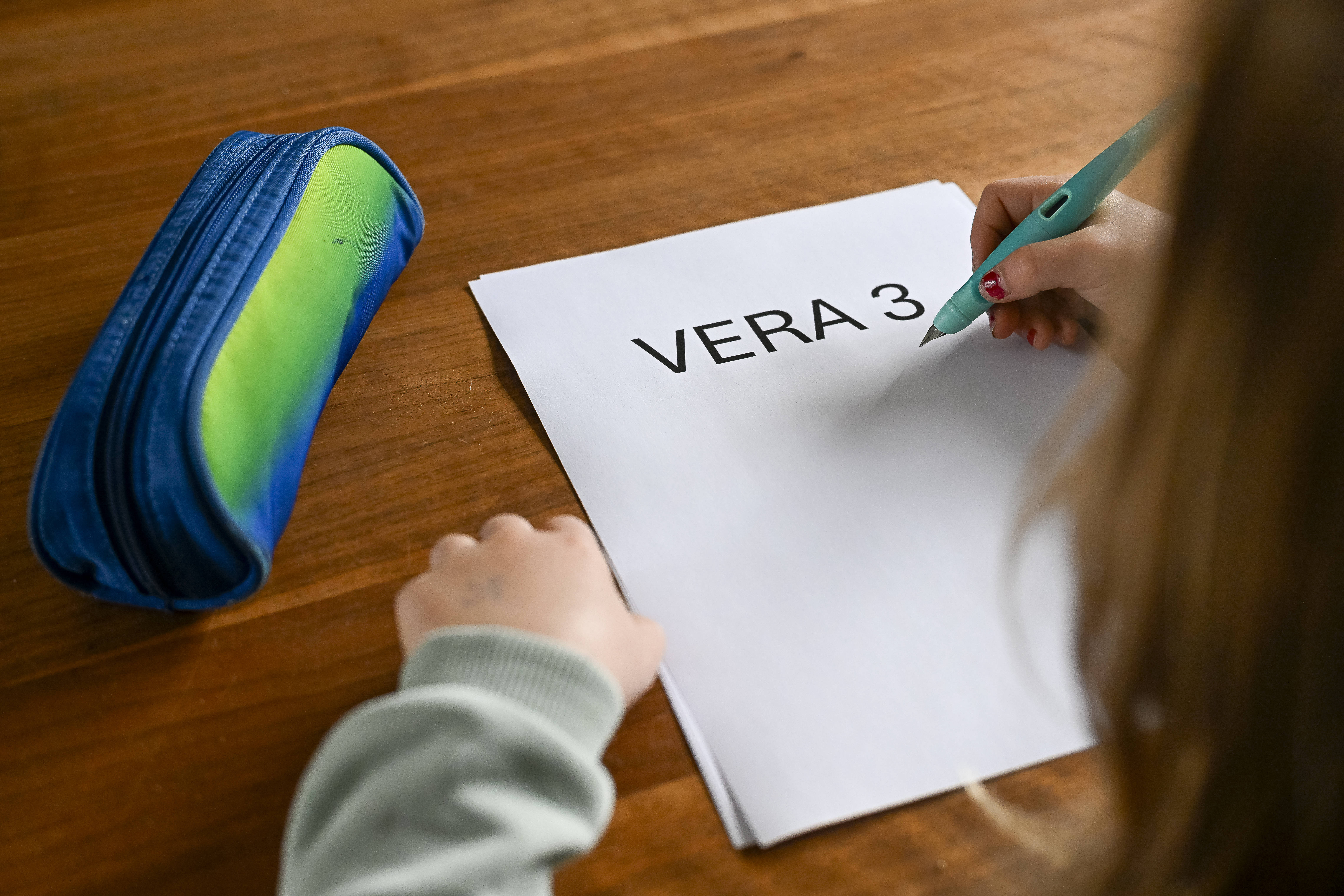 Rückenansicht eines Kindes, das an einem Schreibtisch sitzt. Vor ihm auf dem Tisch ein Blatt, auf dem VERA3 steht. 