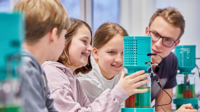 Gruppe von Schülern mit Lehrperson arbeiten an einem Fotobioreaktor, in dem Mikroalgen gezüchtet werden.