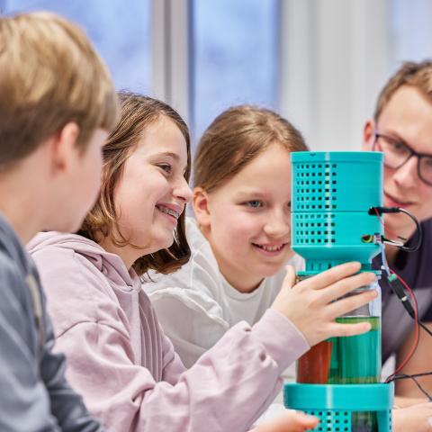 Gruppe von Schülern mit Lehrperson arbeiten an einem Fotobioreaktor, in dem Mikroalgen gezüchtet werden.