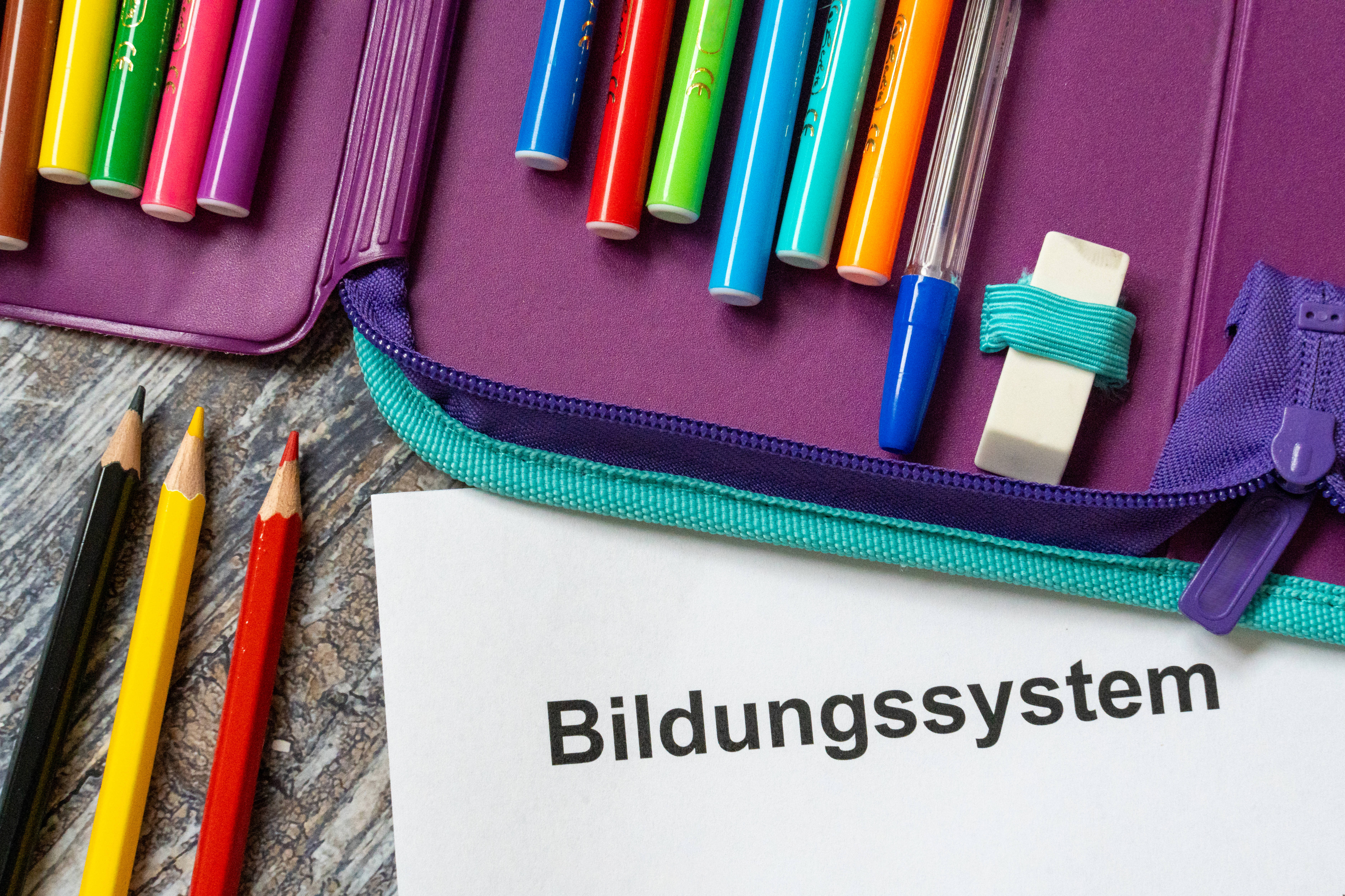 Mäppchen mit Buntstiften auf einem Tisch. Darunter ein Blatt Papier, auf dem "Bildungssystem" steht. 
