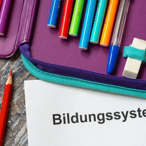 Mäppchen mit Buntstiften auf einem Tisch. Darunter ein Blatt Papier, auf dem "Bildungssystem" steht. 